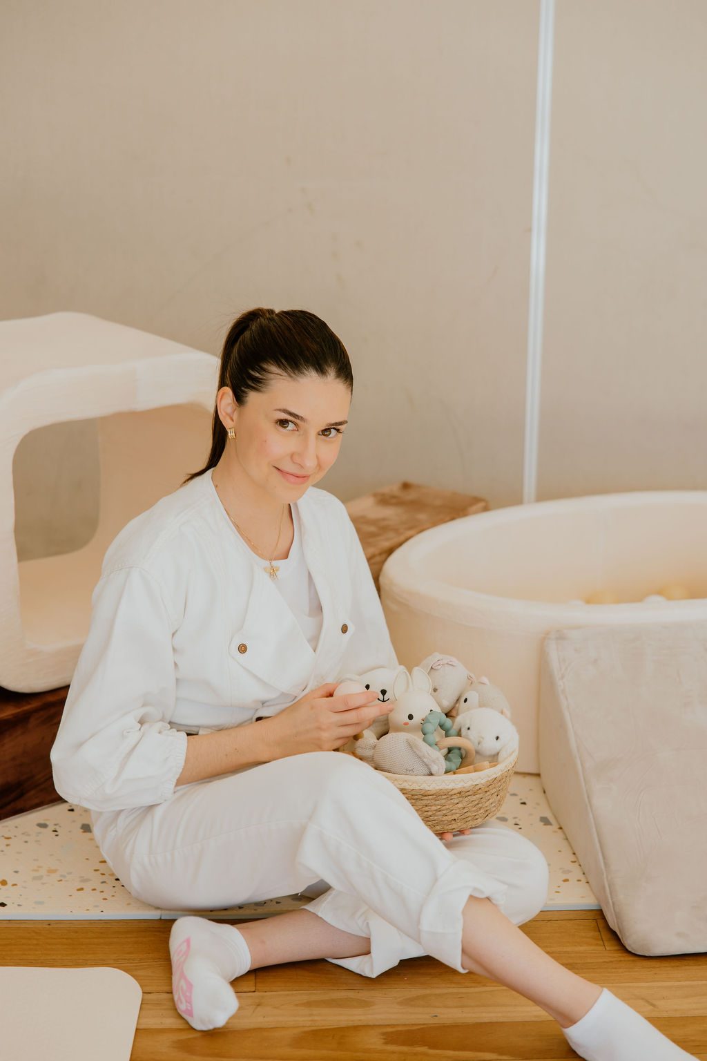 Camila sits on the timber studio floor with a basket of soft knitted toys in her lap, smiling gently, framed between the soft-play blocks and the ball pit