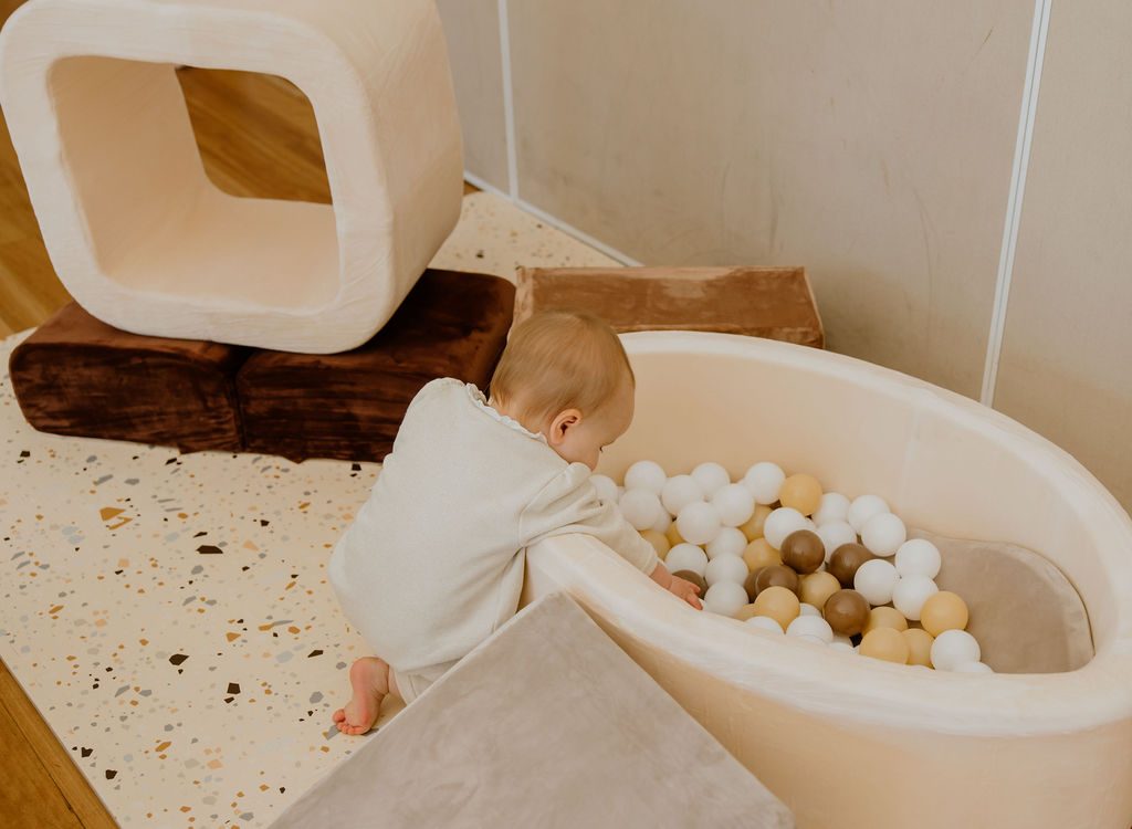 A baby in a cream romper leans over the edge of a soft, neutral-toned ball pit surrounded by foam play blocks