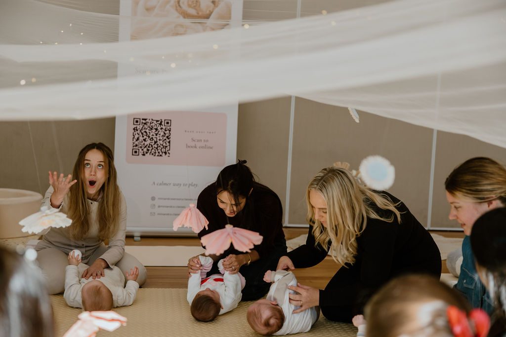 Mums and babies playing together with soft parachute toys under draped fabric at a Mini & Co. sensory class