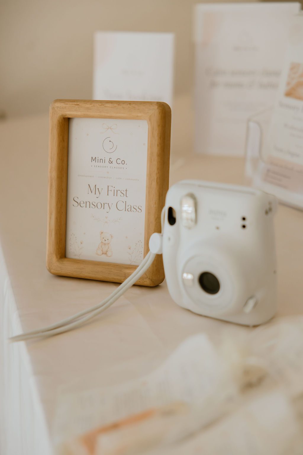 A wooden-framed &lsquo;My First Sensory Class&rsquo; keepsake card beside a white instant camera on a soft cream table
