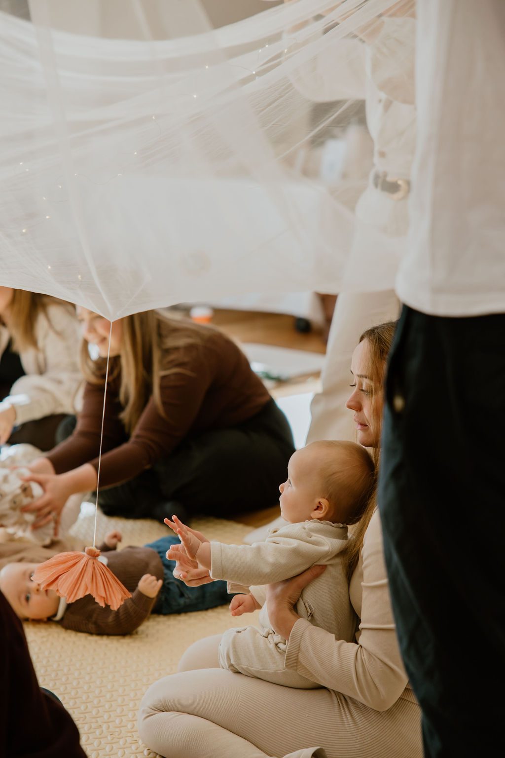 A mum holds her baby as the little one reaches up toward a hanging parachute toy