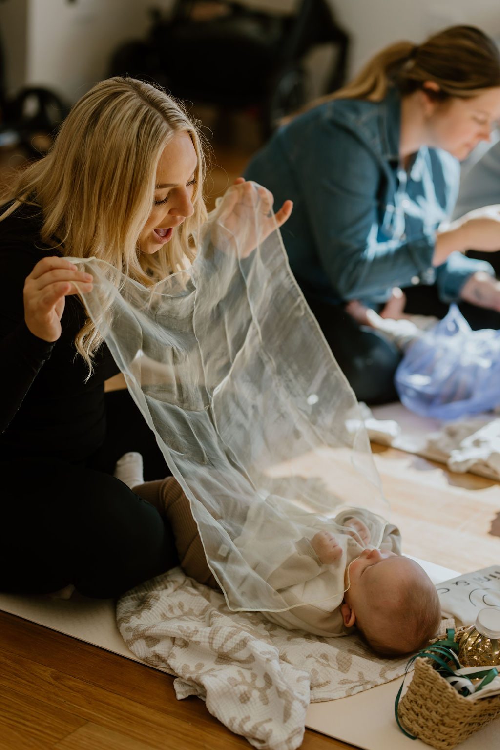 A mum floats a sheer cream play scarf down over her baby, who lies watching, during a Mini & Co. sensory class
