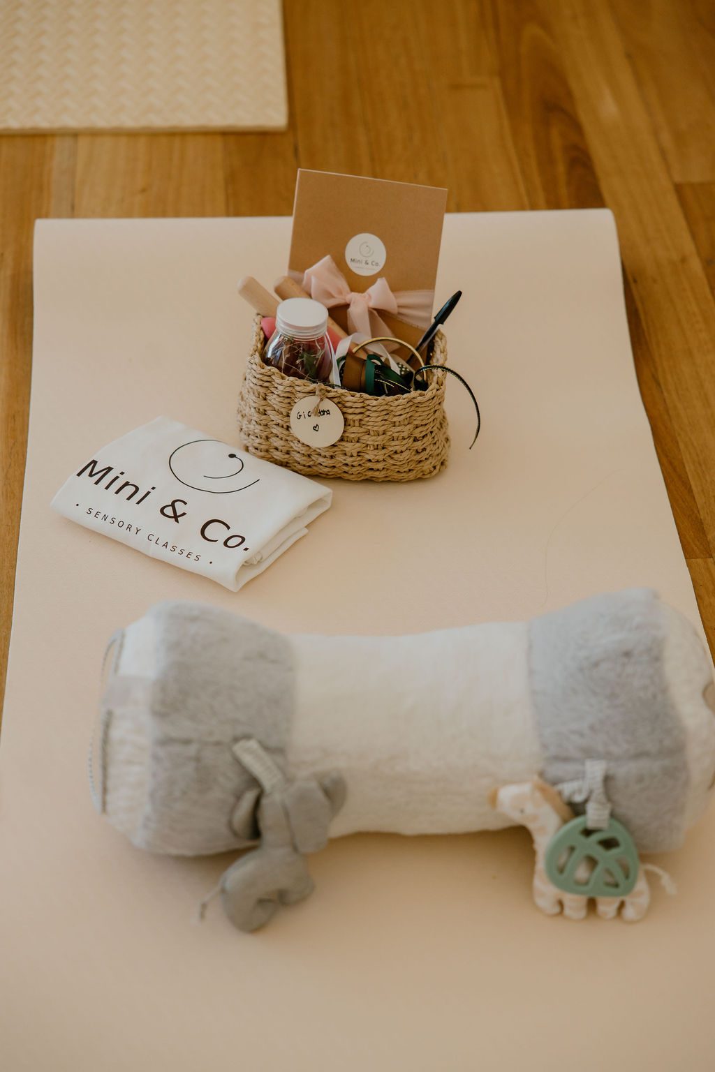 A Mini & Co. welcome basket of sensory tools and a folded branded cloth laid out on a cream play mat